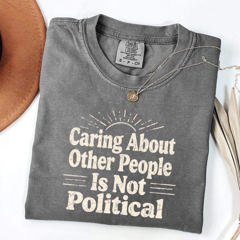 Puede incluir: Una camiseta gris Comfort Colors con el texto "Caring About Other People Is Not Political" en una fuente retro. Tambi&eacute;n se ve un collar plateado con un colgante y un sombrero marr&oacute;n.