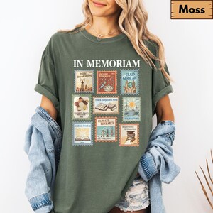 May include: A moss green t-shirt with the words "IN MEMORIAM" above a collection of postage stamp-style graphics. The stamps depict various themes, including "Public Broadcasting," "Vaccine Research," and "Climate Research."