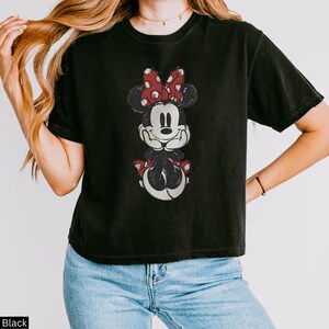 May include: Black t-shirt featuring a distressed graphic of Minnie Mouse. The design shows Minnie with a red bow with white polka dots. The shirt is a crew neck and has short sleeves. The shirt is worn by a person.