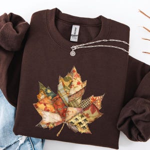 Op de afbeelding: Een bruine sweatshirt met een groot patchwork bladontwerp in herfstkleuren. Een zilveren ketting met een zonnehanger ligt op de trui. De sweatshirt is opgevouwen met een blauwe jeans en een bruine mouw.