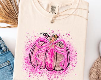 Op de afbeelding: Ivoor Comfort Colors T-shirt met een roze pompoenontwerp, een roze lint en borstkanker bewustzijns linten. De pompoen heeft een glittertextuur en roze verfspatten. Een gouden ketting is over het shirt gedrapeerd.