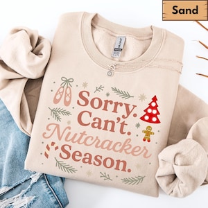 Puede incluir: Una sudadera de color arena con el texto "Sorry. Can't. Nutcracker Season." en una fuente festiva. El diseño incluye zapatillas de ballet, un árbol de Navidad y un hombre de jengibre. La sudadera es de material suave.