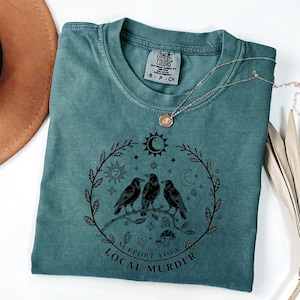 May include: Teal Comfort Colors t-shirt with a black graphic of three crows perched on a branch, surrounded by a wreath of leaves, sun, moon, and stars. The text "Support Your Local Murder" is below the crows. A gold necklace is draped across the shirt.