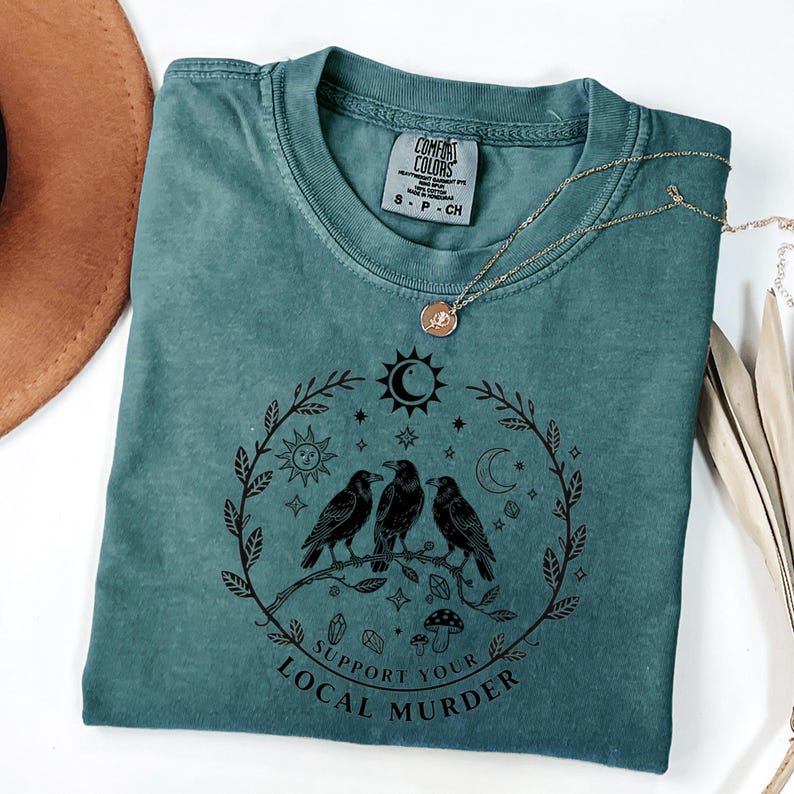May include: Teal Comfort Colors t-shirt with a black graphic of three crows perched on a branch, surrounded by a wreath of leaves, sun, moon, and stars. The text "Support Your Local Murder" is below the crows. A gold necklace is draped across the shirt.