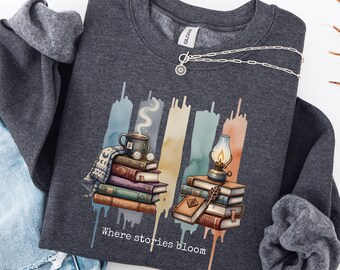 May include: Dark heather grey sweatshirt featuring a graphic of books, a steaming mug, and a lantern. The design includes watercolour-style vertical stripes and the text "Where stories bloom". A silver necklace with a sun pendant is also visible.