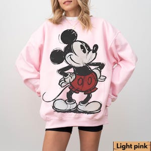 Puede incluir: Sudadera rosa claro con un gr&aacute;fico de Mickey Mouse en negro, rojo y blanco. La parte superior de manga larga tiene cuello redondo y un ajuste relajado. El texto "Light pink" est&aacute; en la parte inferior de la imagen.
