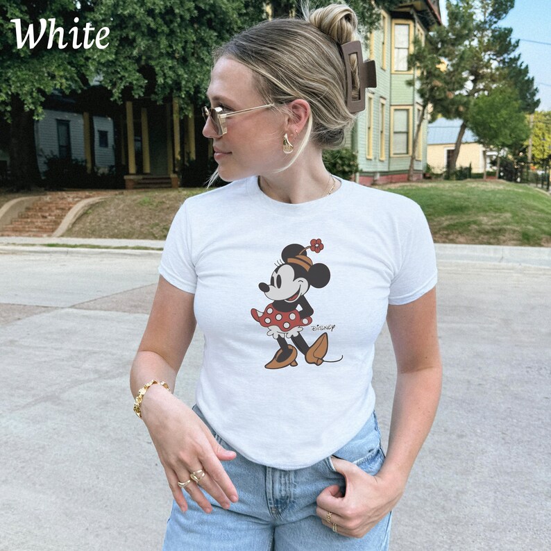May include: White short-sleeve t-shirt featuring a Minnie Mouse graphic. The cartoon character is depicted in a red and white polka-dot dress and brown shoes. The word "White" is printed in the upper left corner of the image.