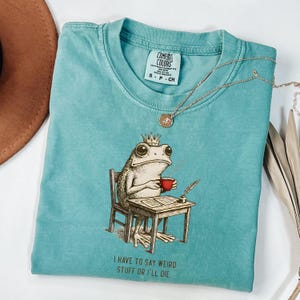 K&ouml;nnte beinhalten: Ein t&uuml;rkisfarbenes Comfort Colors T-Shirt mit einer skurrilen Frosch-Illustration. Der Frosch, mit Krone, sitzt an einem Schreibtisch und h&auml;lt eine rote Tasse. Der Text "I HAVE TO SAY WEIRD STUFF OR I'LL DIE" ist darunter gedruckt. Eine goldene Halskette liegt auf dem Shirt.