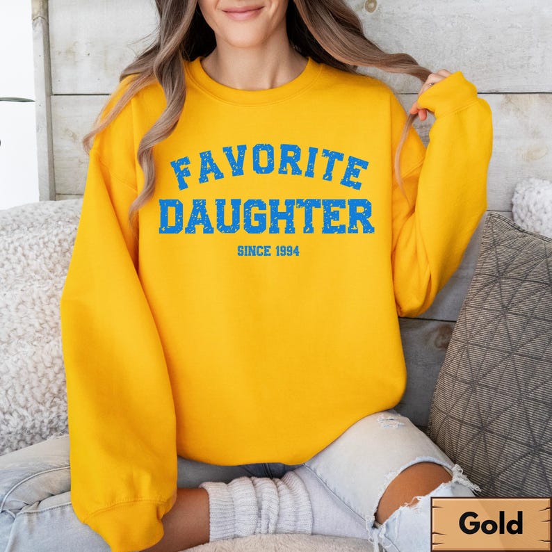 Puede incluir: Una sudadera dorada con el texto azul "FAVORITE DAUGHTER" y "SINCE 1994". La sudadera tiene mangas largas y cuello redondo. La imagen muestra a una persona que lleva la sudadera.