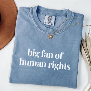 Pode incluir: Uma camiseta azul Comfort Colors com o texto "big fan of human rights" em branco. Um colar de ouro com um pingente repousa sobre a camiseta. A camiseta é tamanho pequeno.