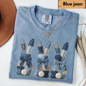 May include: A light blue t-shirt with three bunny appliqués. Each bunny is made of patchwork denim with a bow and a white pom-pom tail. The shirt has the text "Blue jean" at the top.