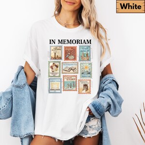 May include: White t-shirt with the text "IN MEMORIAM" above a collection of postage stamp designs. The stamps feature illustrations and text related to various topics, including "Public Broadcasting" and "Climate Research."