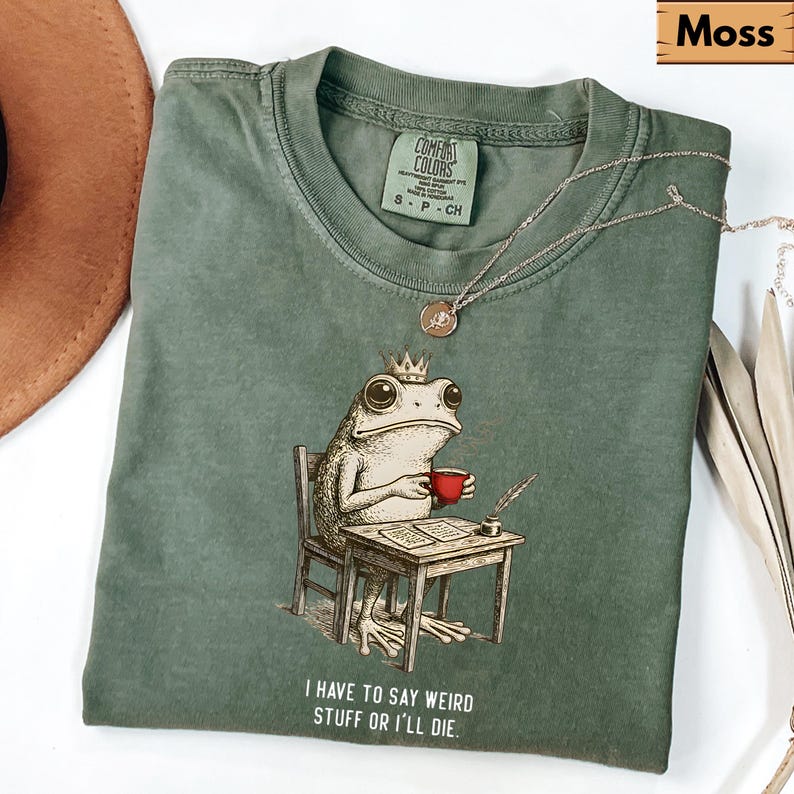 K&ouml;nnte beinhalten: Moosgr&uuml;nes T-Shirt mit einer humorvollen Illustration einer Froschk&ouml;nigin, die eine rote Tasse h&auml;lt und an einem Schreibtisch sitzt. Der Text "I HAVE TO SAY WEIRD STUFF OR I'LL DIE" ist darunter gedruckt. Eine silberne Halskette liegt auf dem T-Shirt.
