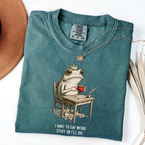 K&ouml;nnte beinhalten: T&uuml;rkisfarbenes Comfort Colors T-Shirt mit einer Illustration einer gekr&ouml;nten Froschfigur, die eine rote Tasse h&auml;lt und an einem Schreibtisch sitzt. Der Text "I HAVE TO SAY WEIRD STUFF OR I'LL DIE" ist darunter gedruckt. Eine goldene Halskette liegt auf dem Shirt.