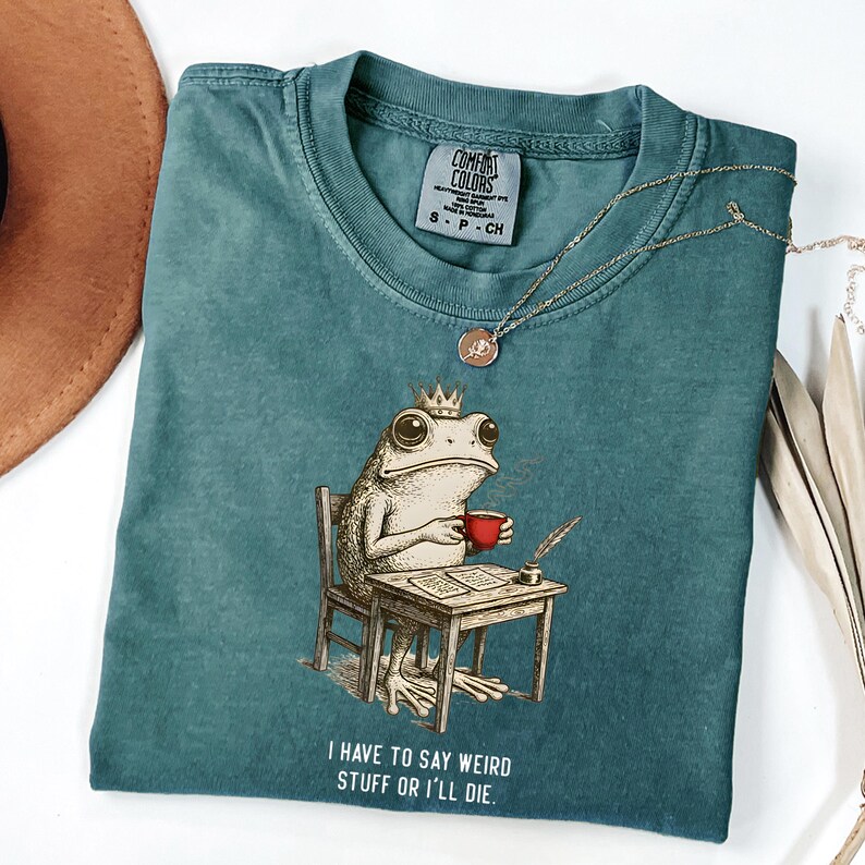 K&ouml;nnte beinhalten: T&uuml;rkisfarbenes Comfort Colors T-Shirt mit einer Illustration einer gekr&ouml;nten Froschfigur, die eine rote Tasse h&auml;lt und an einem Schreibtisch sitzt. Der Text "I HAVE TO SAY WEIRD STUFF OR I'LL DIE" ist darunter gedruckt. Eine goldene Halskette liegt auf dem Shirt.