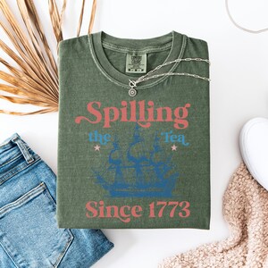 May include: Green t-shirt with a vintage-style graphic that says "Spilling the Tea Since 1773". The graphic features a blue ship with red and blue text.