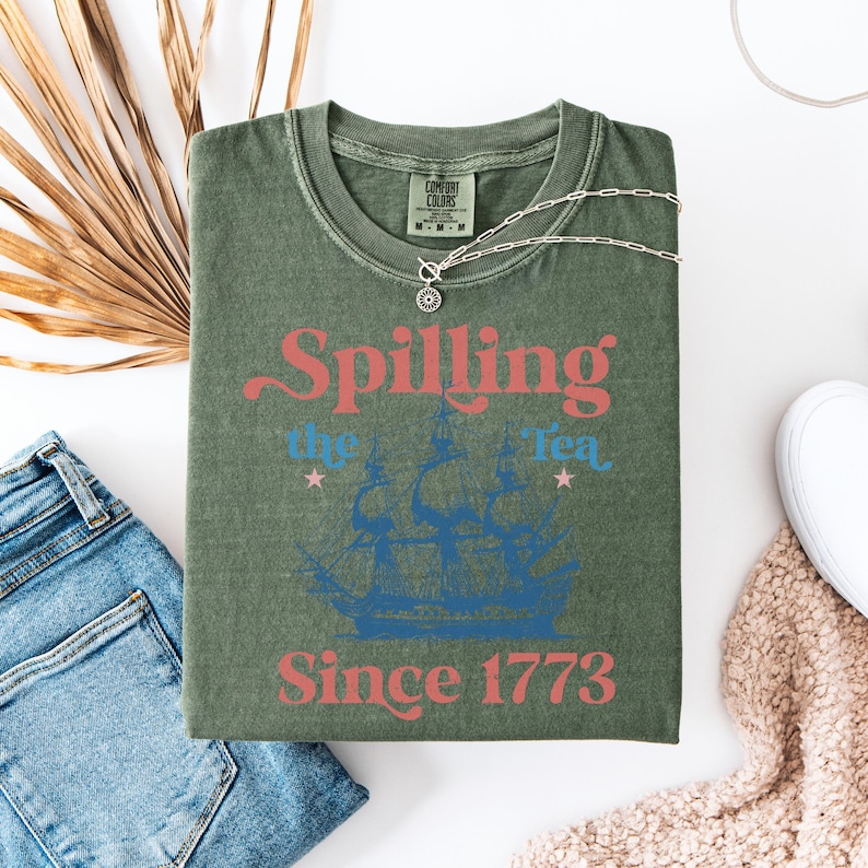 May include: Green t-shirt with a vintage-style graphic that says "Spilling the Tea Since 1773". The graphic features a blue ship with red and blue text.
