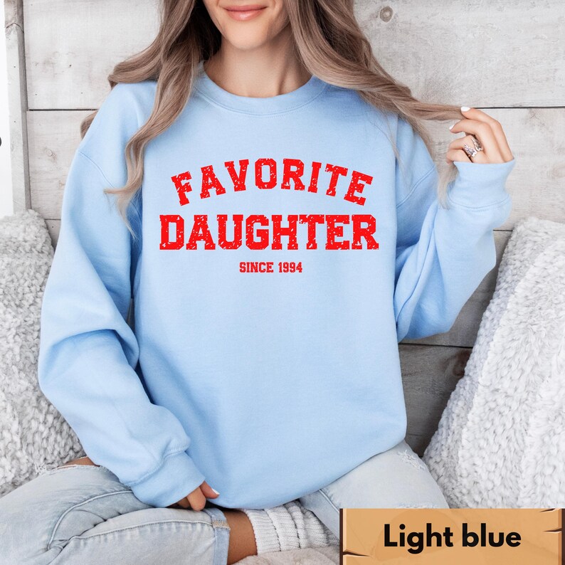 Puede incluir: Sudadera azul claro con la frase "FAVORITE DAUGHTER" en rojo, con letras desgastadas, y "SINCE 1994" debajo. La sudadera es de color azul claro liso.