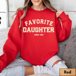 Puede incluir: Una sudadera roja con las palabras "FAVORITE DAUGHTER" en una fuente desgastada de color blanquecino. Debajo del texto est&aacute; "SINCE 1994". La sudadera es de color rojo liso y tiene cuello redondo.
