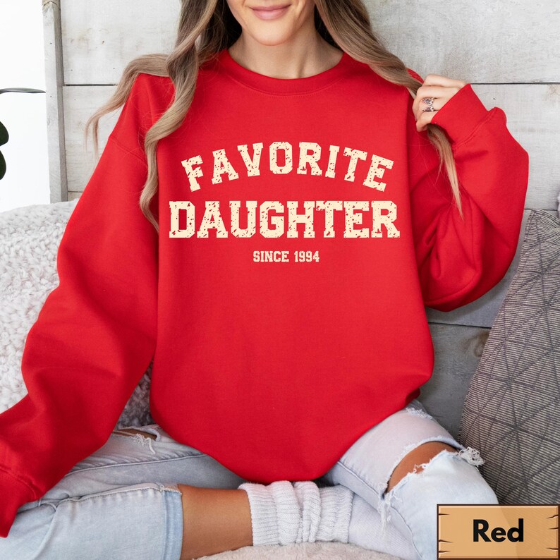 Puede incluir: Una sudadera roja con las palabras "FAVORITE DAUGHTER" en una fuente desgastada de color blanquecino. Debajo del texto est&aacute; "SINCE 1994". La sudadera es de color rojo liso y tiene cuello redondo.