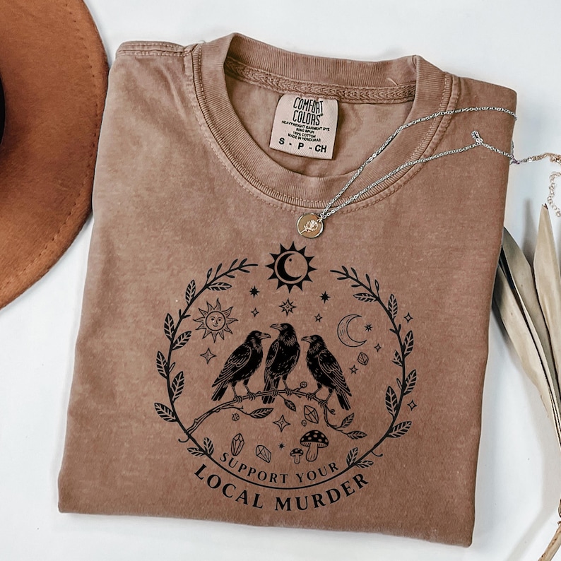 May include: A brown Comfort Colors t-shirt with a round neck and short sleeves. The shirt features a black graphic of three crows perched on a branch, surrounded by celestial elements and the text "Support Your Local Murder."