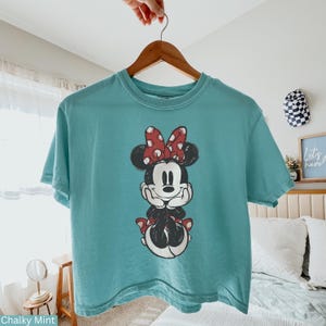 May include: A teal-colored t-shirt featuring a graphic of Minnie Mouse. Minnie Mouse is depicted in black, white, and red, with a polka-dot bow. The shirt is hanging on a wooden hanger.