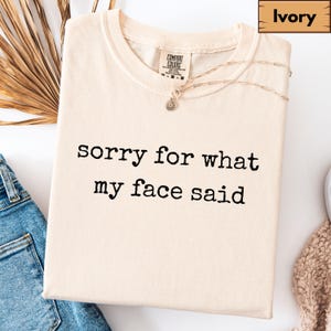 May include: Ivory-colored t-shirt with the text "sorry for what my face said" in black. The shirt has a crew neck and short sleeves. A gold necklace with a small pendant is draped across the shirt. The shirt is folded on a white surface.