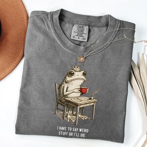 K&ouml;nnte beinhalten: Ein graues Comfort Colors T-Shirt mit einer skurrilen Illustration einer Froschk&ouml;nigin, die an einem Schreibtisch sitzt und eine rote Tasse h&auml;lt. Der Text "I HAVE TO SAY WEIRD STUFF OR I'LL DIE" ist darunter gedruckt. Eine silberne Halskette liegt auf dem Shirt.