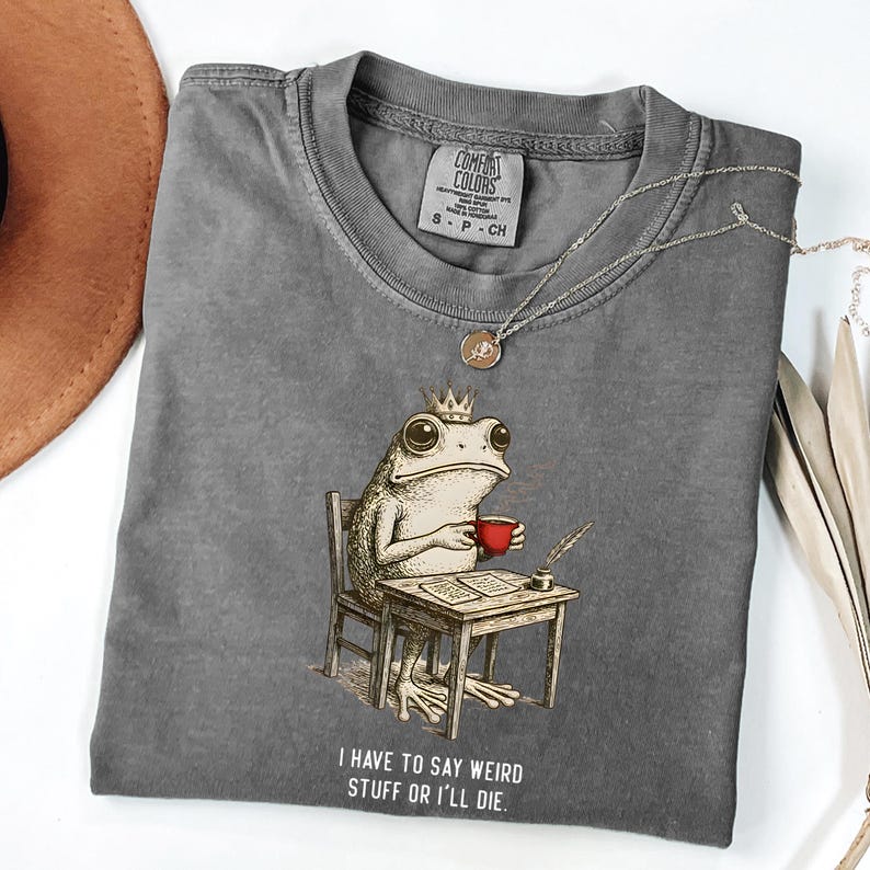 K&ouml;nnte beinhalten: Ein graues Comfort Colors T-Shirt mit einer skurrilen Illustration einer Froschk&ouml;nigin, die an einem Schreibtisch sitzt und eine rote Tasse h&auml;lt. Der Text "I HAVE TO SAY WEIRD STUFF OR I'LL DIE" ist darunter gedruckt. Eine silberne Halskette liegt auf dem Shirt.