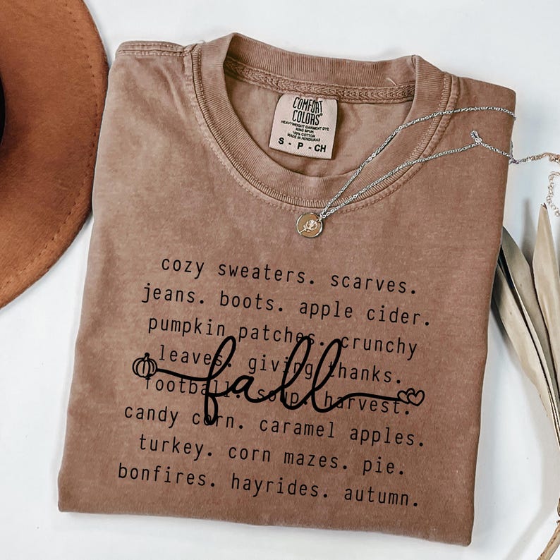 K&ouml;nnte beinhalten: Ein braunes Comfort Colors T-Shirt mit dem Wort "fall" in Schreibschrift. Das Shirt listet herbstliche W&ouml;rter wie "cozy sweaters", "pumpkin patches" und "hayrides" auf. Eine silberne Halskette mit einem kleinen Anh&auml;nger liegt auf dem Shirt.