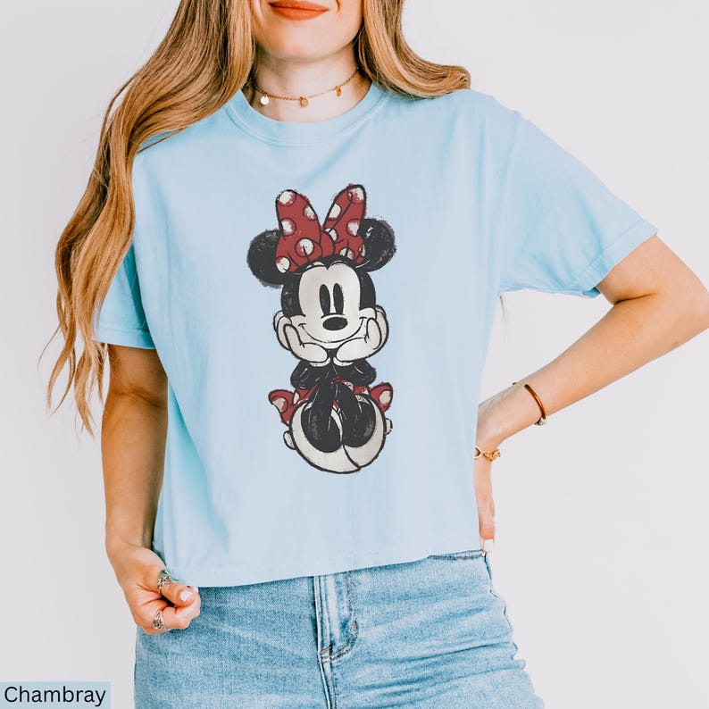 May include: Light blue t-shirt featuring a distressed graphic of Minnie Mouse with a red polka dot bow. The cartoon character is smiling and seated. The shirt is made of a soft material and has short sleeves. The word "Chambray" is printed at the bottom left.