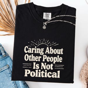Puede incluir: Camiseta negra con el texto "Caring About Other People Is Not Political" en blanco, con una fuente de estilo vintage. Un collar plateado con un peque&ntilde;o colgante est&aacute; drapeado sobre la camiseta. La camiseta est&aacute; doblada y colocada sobre una superficie blanca.