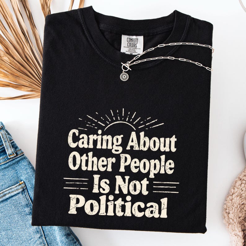Puede incluir: Camiseta negra con el texto "Caring About Other People Is Not Political" en blanco, con una fuente de estilo vintage. Un collar plateado con un peque&ntilde;o colgante est&aacute; drapeado sobre la camiseta. La camiseta est&aacute; doblada y colocada sobre una superficie blanca.