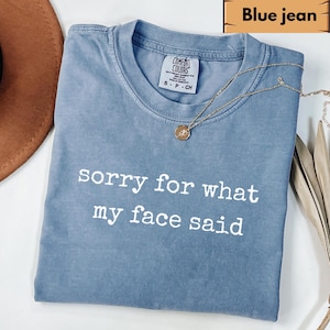 May include: Blue jean colored t-shirt with the text "sorry for what my face said" in white. The shirt has a crew neck and short sleeves. A gold necklace with a pendant is draped across the shirt.