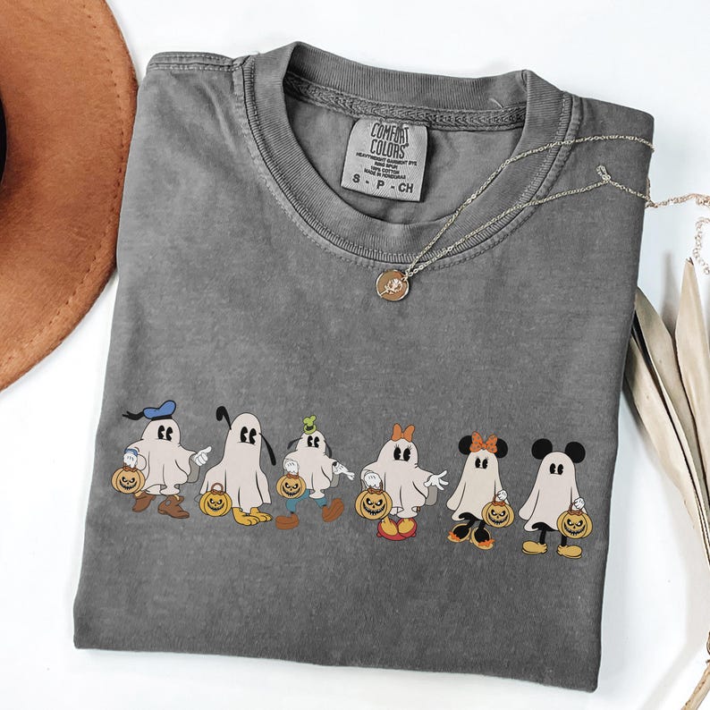 Op de afbeelding: Een grijs Comfort Colors T-shirt met een rij cartoongeesten, elk met een pompoen. De geesten zijn gestyled naar populaire stripfiguren. Een zilveren ketting met een hanger ligt op het shirt.