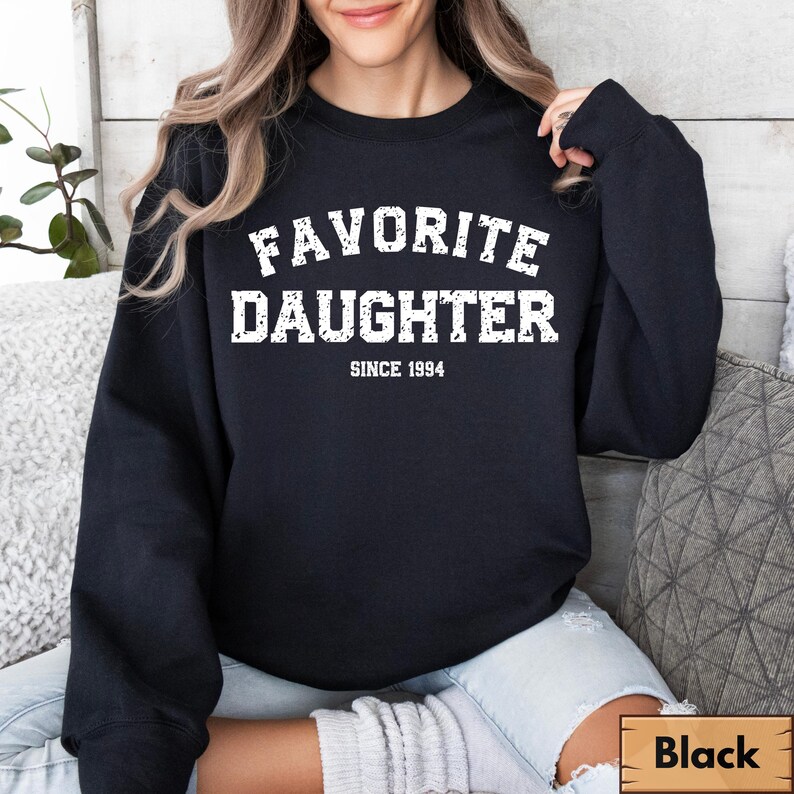 Puede incluir: Sudadera negra con el texto blanco "FAVORITE DAUGHTER" y "SINCE 1994". Un letrero de madera en la esquina inferior derecha dice "Black". La sudadera es de estilo casual.