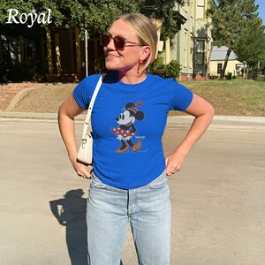 May include: Royal blue t-shirt featuring a Minnie Mouse graphic. The cartoon character is wearing a red polka dot dress and brown shoes. The word "Royal" is printed in white in the upper left corner. The shirt is paired with light blue jeans.