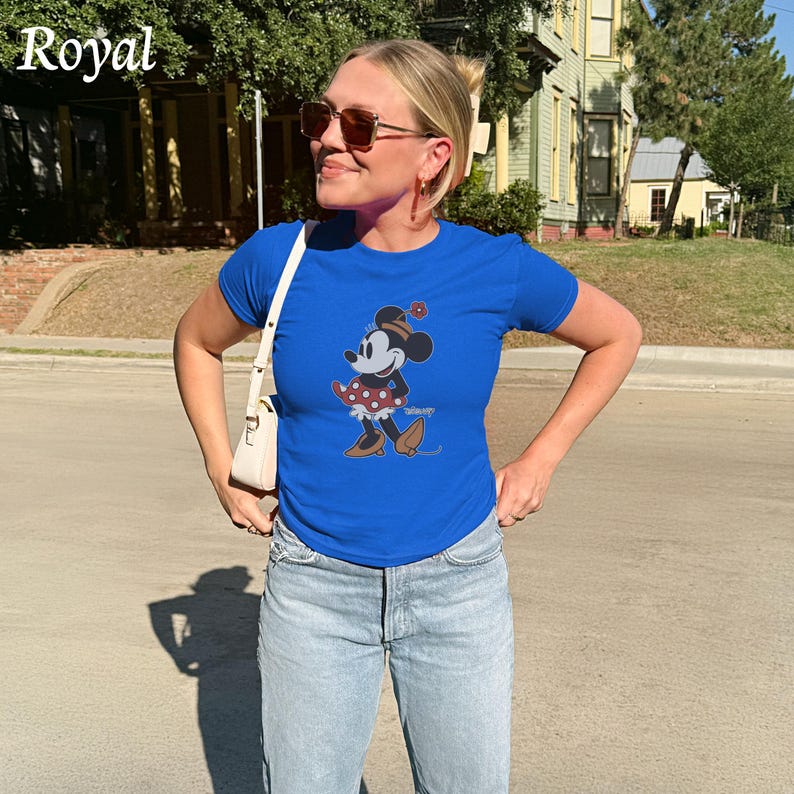 May include: Royal blue t-shirt featuring a Minnie Mouse graphic. The cartoon character is wearing a red polka dot dress and brown shoes. The word "Royal" is printed in white in the upper left corner. The shirt is paired with light blue jeans.