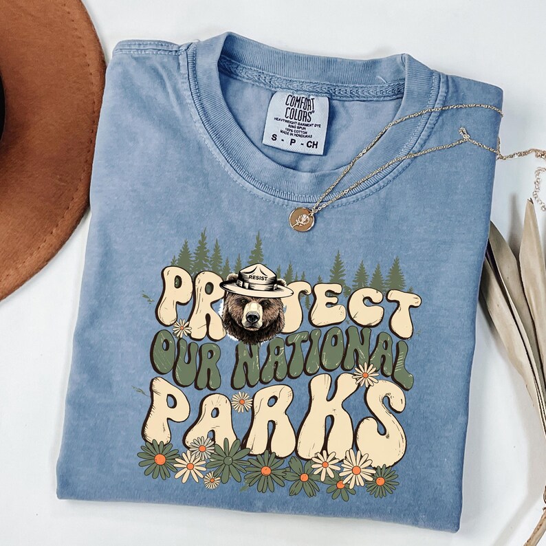 Puede incluir: Una camiseta azul Comfort Colors con el texto "PROTECT OUR NATIONAL PARKS" en una fuente retro. El dise&ntilde;o incluye un oso con sombrero de guardabosques, &aacute;rboles y margaritas. Un collar dorado est&aacute; drapeado sobre la camisa.