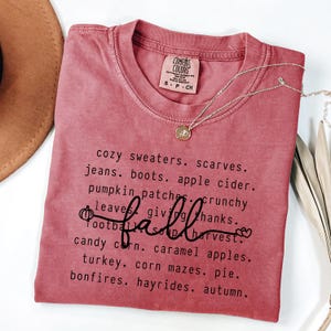 K&ouml;nnte beinhalten: Ein staubrosa Comfort Colors T-Shirt mit dem Wort "fall" in Schreibschrift und einer Liste von W&ouml;rtern zum Thema Herbst. Eine goldene Halskette mit Anh&auml;nger ist auf dem T-Shirt drapiert.