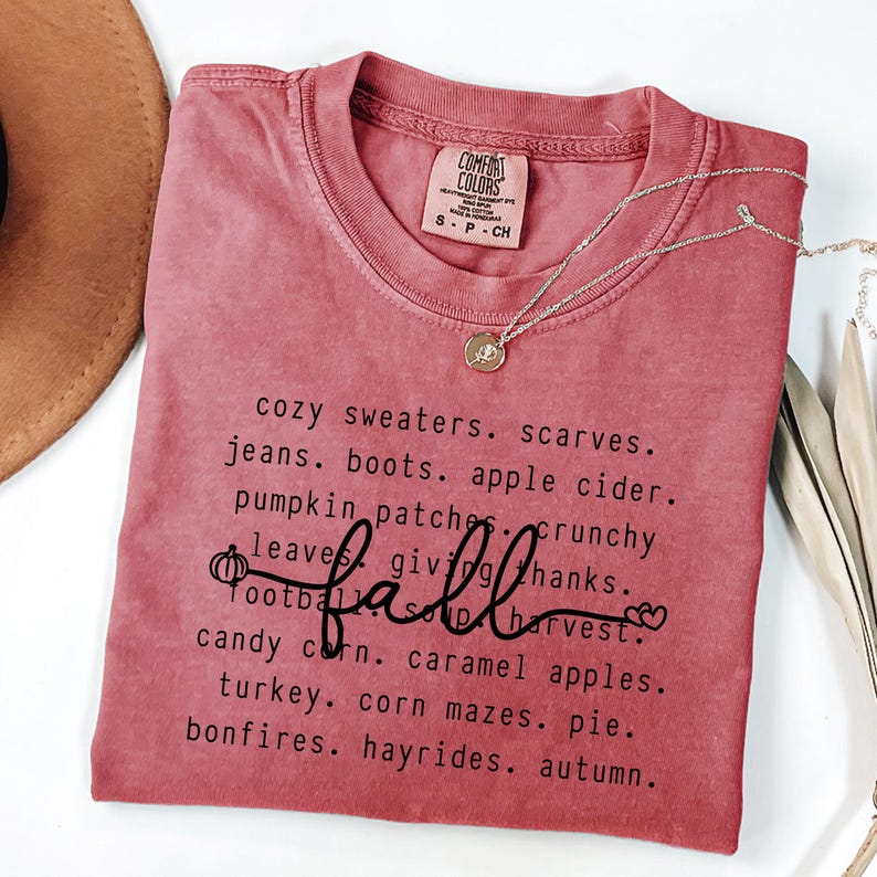 K&ouml;nnte beinhalten: Ein staubrosa Comfort Colors T-Shirt mit dem Wort "fall" in Schreibschrift und einer Liste von W&ouml;rtern zum Thema Herbst. Eine goldene Halskette mit Anh&auml;nger ist auf dem T-Shirt drapiert.