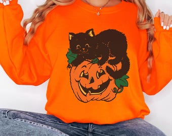May include: A bright orange long-sleeve sweatshirt featuring a graphic print of a black kitten sitting on top of a carved jack-o'-lantern pumpkin with green leaves. The sweatshirt has a crew neckline and long sleeves with ribbed cuffs.