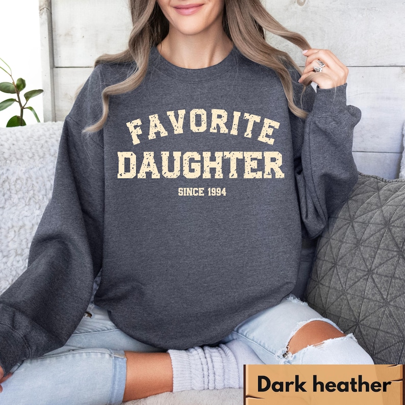 Puede incluir: Sudadera gris oscuro con el texto "FAVORITE DAUGHTER SINCE 1994" en una fuente desgastada de color blanquecino. La sudadera de cuello redondo est&aacute; hecha de un material suave. La imagen muestra la sudadera en una persona.