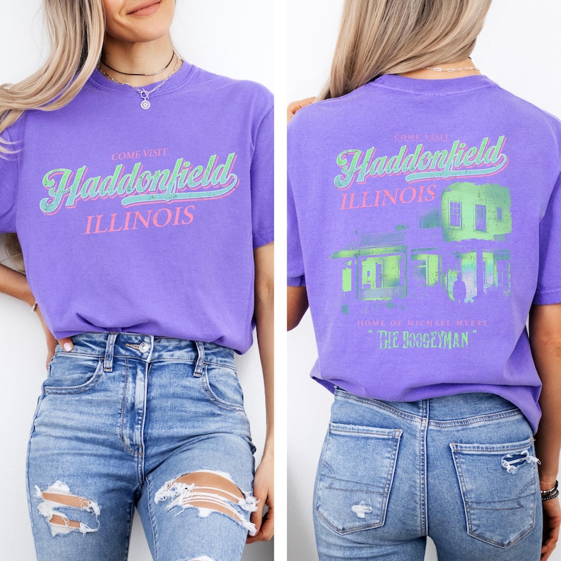 May include: A purple t-shirt with the text "COME VISIT Haddonfield ILLINOIS" in a retro font on the front. The back of the shirt features a green and blue graphic of a house and the text "HOME OF MICHAEL MYERS THE BOOGEYMAN."