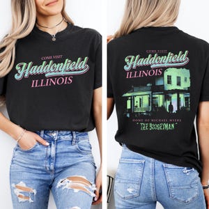 May include: Black t-shirt with the words "Come Visit Haddonfield Illinois" in a retro font on the front. The back of the shirt features a house graphic and the text "Home of Michael Myers" and "The Boogeyman."