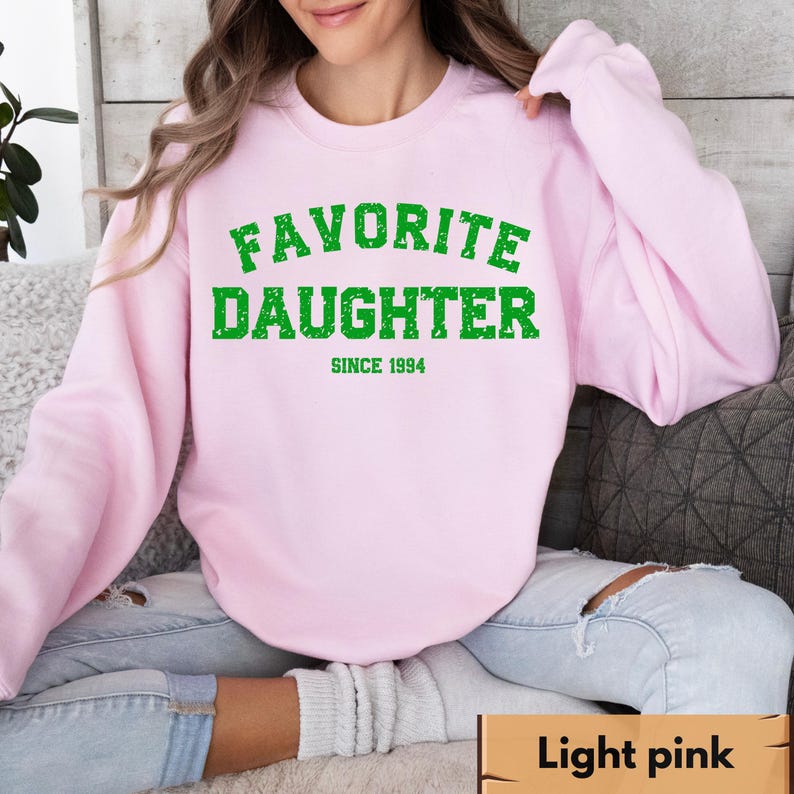 Puede incluir: Sudadera rosa claro con el texto "FAVORITE DAUGHTER" en verde, con "SINCE 1994" debajo. La sudadera es de color rosa claro liso. La modelo lleva vaqueros azules.