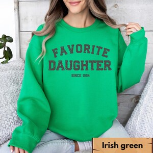 Puede incluir: Sudadera verde irland&eacute;s brillante con el texto "FAVORITE DAUGHTER SINCE 1994" en gris oscuro desgastado. La sudadera de cuello redondo tiene mangas largas. El fondo es una superficie clara y texturizada.