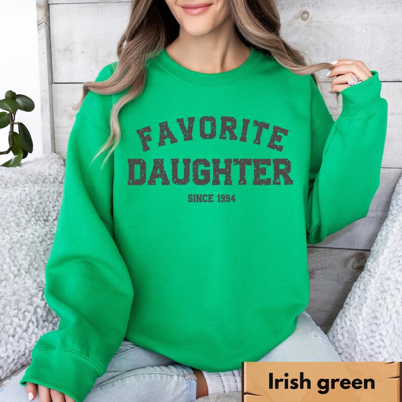 Puede incluir: Sudadera verde irland&eacute;s brillante con el texto "FAVORITE DAUGHTER SINCE 1994" en gris oscuro desgastado. La sudadera de cuello redondo tiene mangas largas. El fondo es una superficie clara y texturizada.