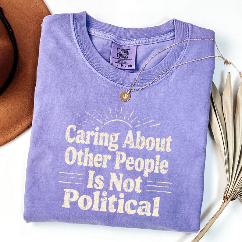 Puede incluir: Una camiseta de color lila con el texto "Caring About Other People Is Not Political" en una fuente vintage. Un collar de oro con un colgante descansa sobre la camiseta. La camiseta es de Comfort Colors.