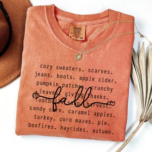 K&ouml;nnte beinhalten: Orangenes Comfort Colors T-Shirt mit einer Liste von W&ouml;rtern zum Thema Herbst in Schwarz, darunter das Wort "fall" in Schreibschrift. Eine goldene Halskette mit Anh&auml;nger liegt auf dem T-Shirt. Das T-Shirt ist auf einer wei&szlig;en Oberfl&auml;che gefaltet.